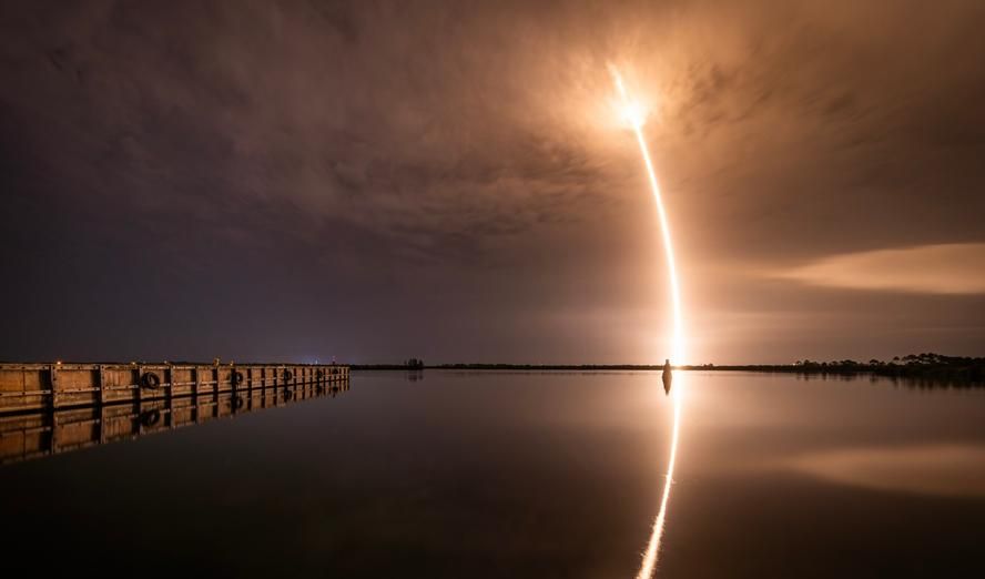Lång exponering av en raketuppskjutning som reflekteras i vattnet på natten.